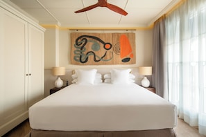 Een groot bed met witte lakens, twee nachtkastjes met lampen, een hangend abstract kunstwerk en een houten plafondventilator.