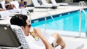 Un homme se détend sur une chaise longue au bord d’une piscine, lisant un livre et tenant un verre à la main.