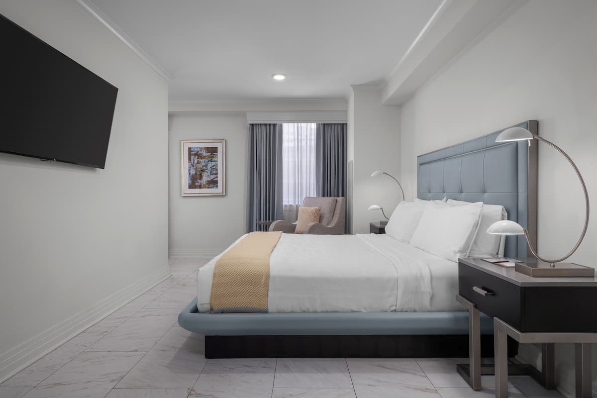 A modern hotel room with a large bed, a bedside table with a lamp, a flat-screen TV mounted on the wall, and a window with curtains.