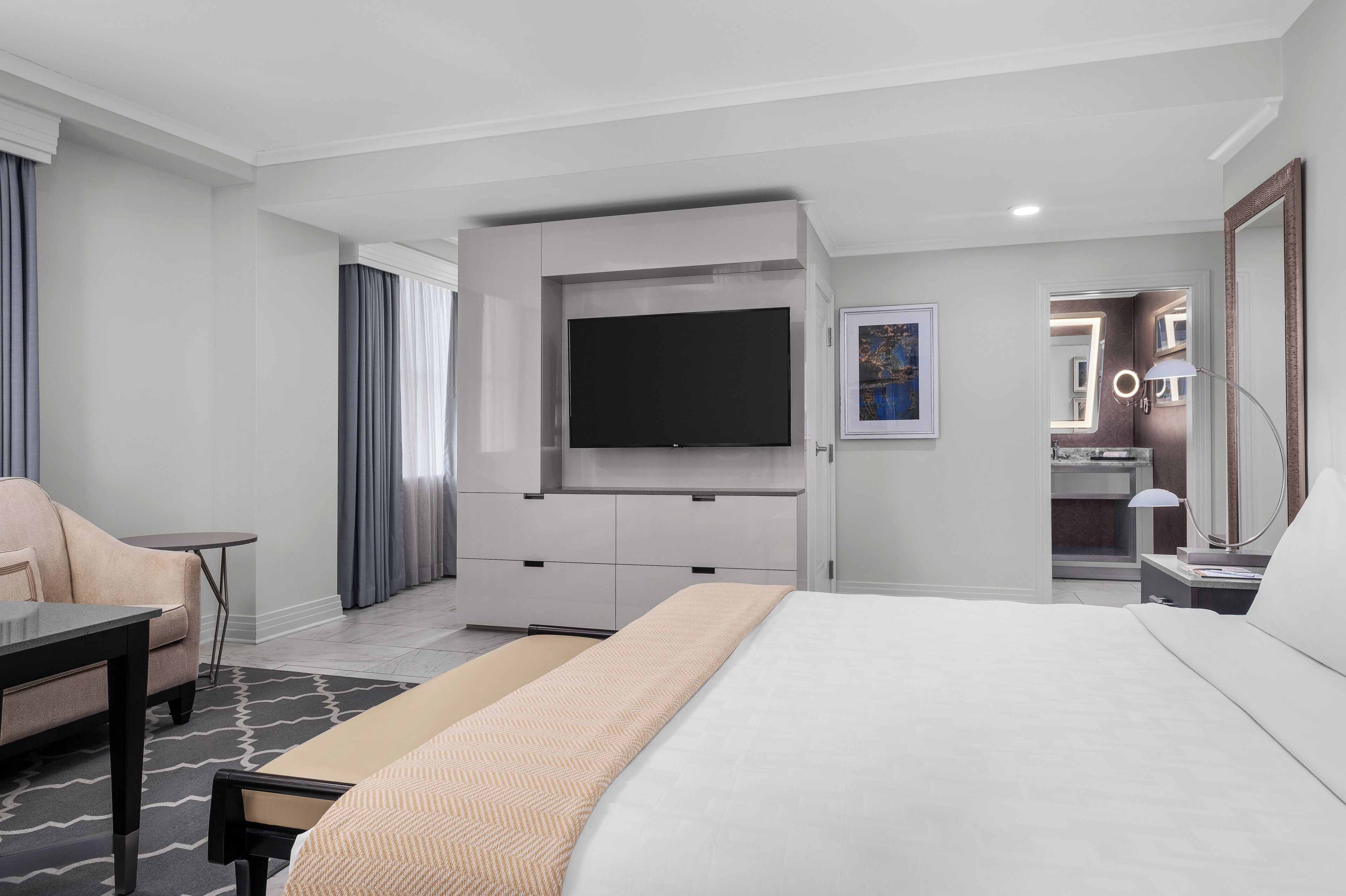 A modern hotel room with a large bed, a flat-screen TV mounted on the wall, a desk with a chair, and a small table.