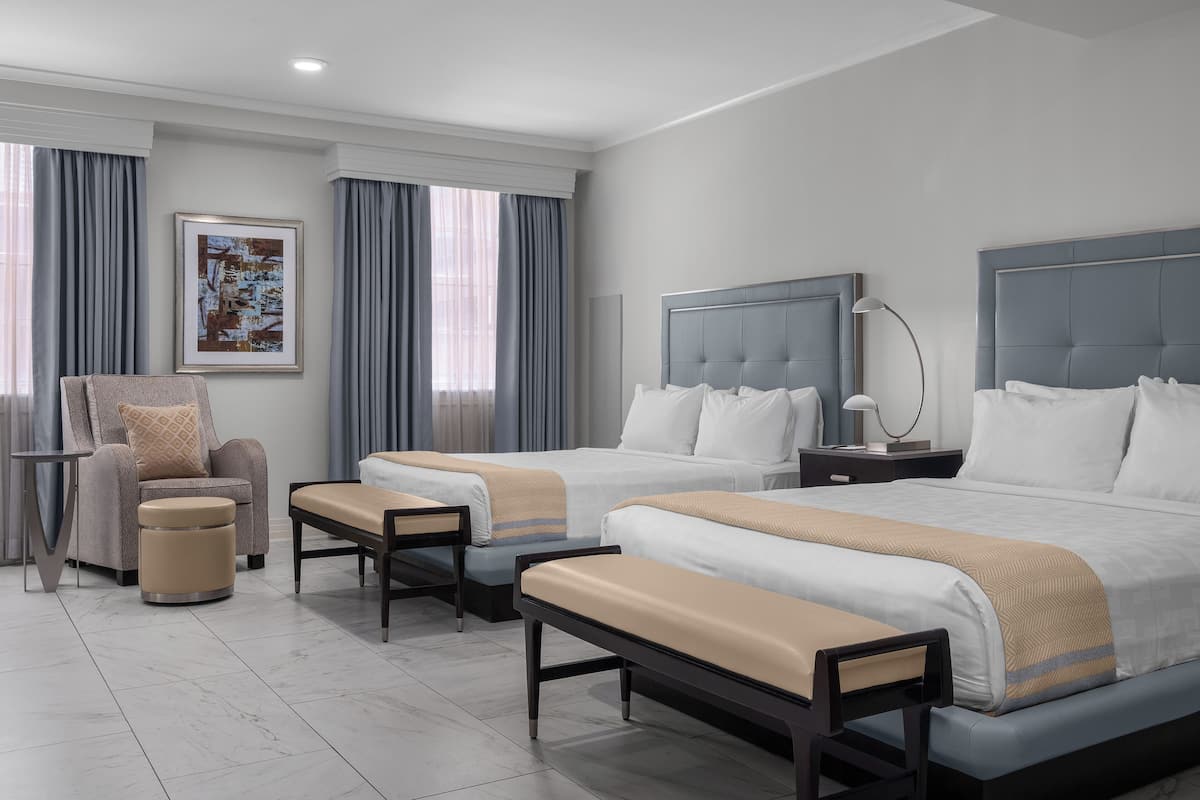 A hotel room with two beds, a chair, a bench, a side table, and a framed picture on the wall.