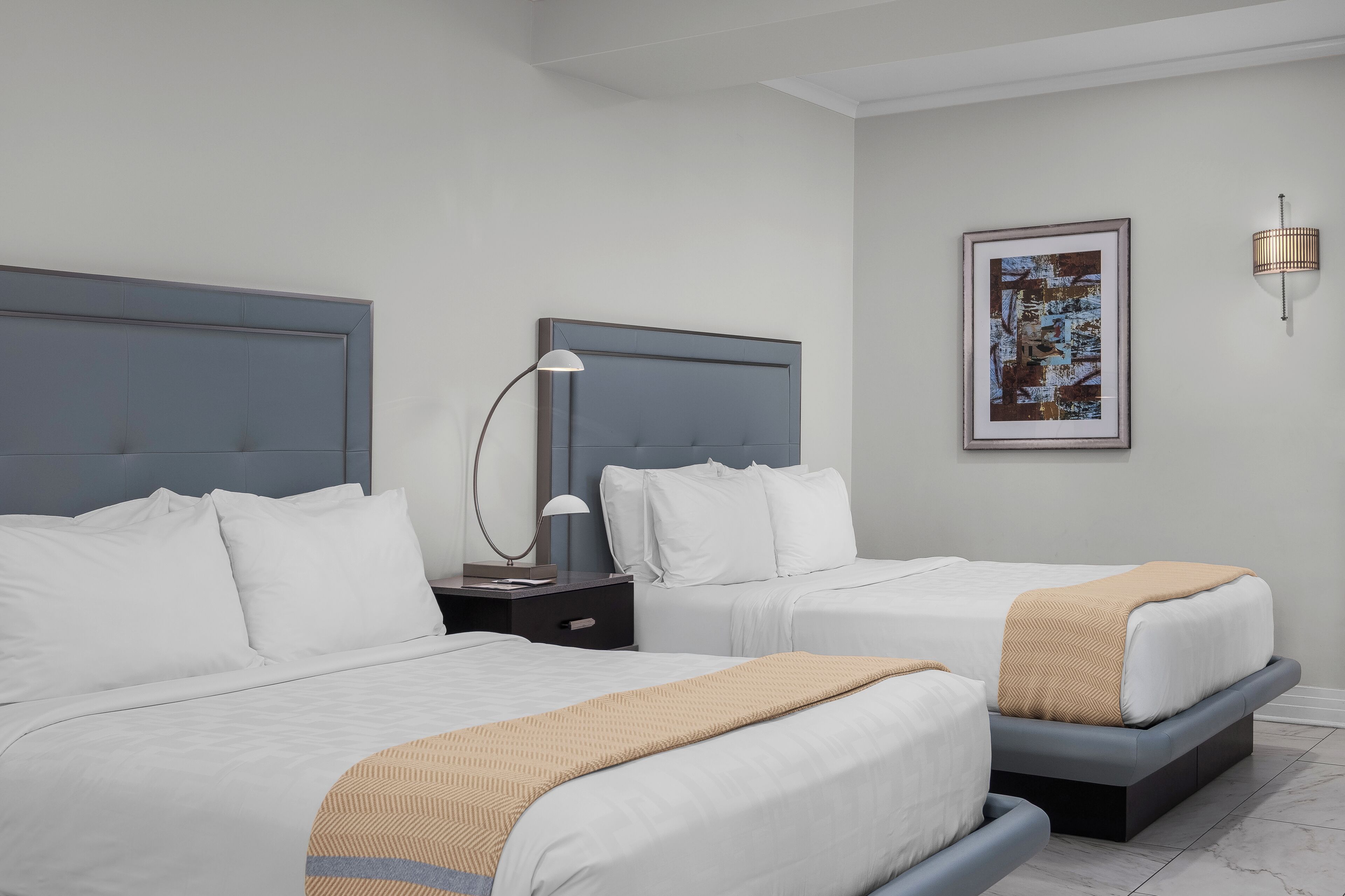 A hotel room with two beds, a nightstand with a lamp, a framed picture on the wall, and a modern light fixture.
