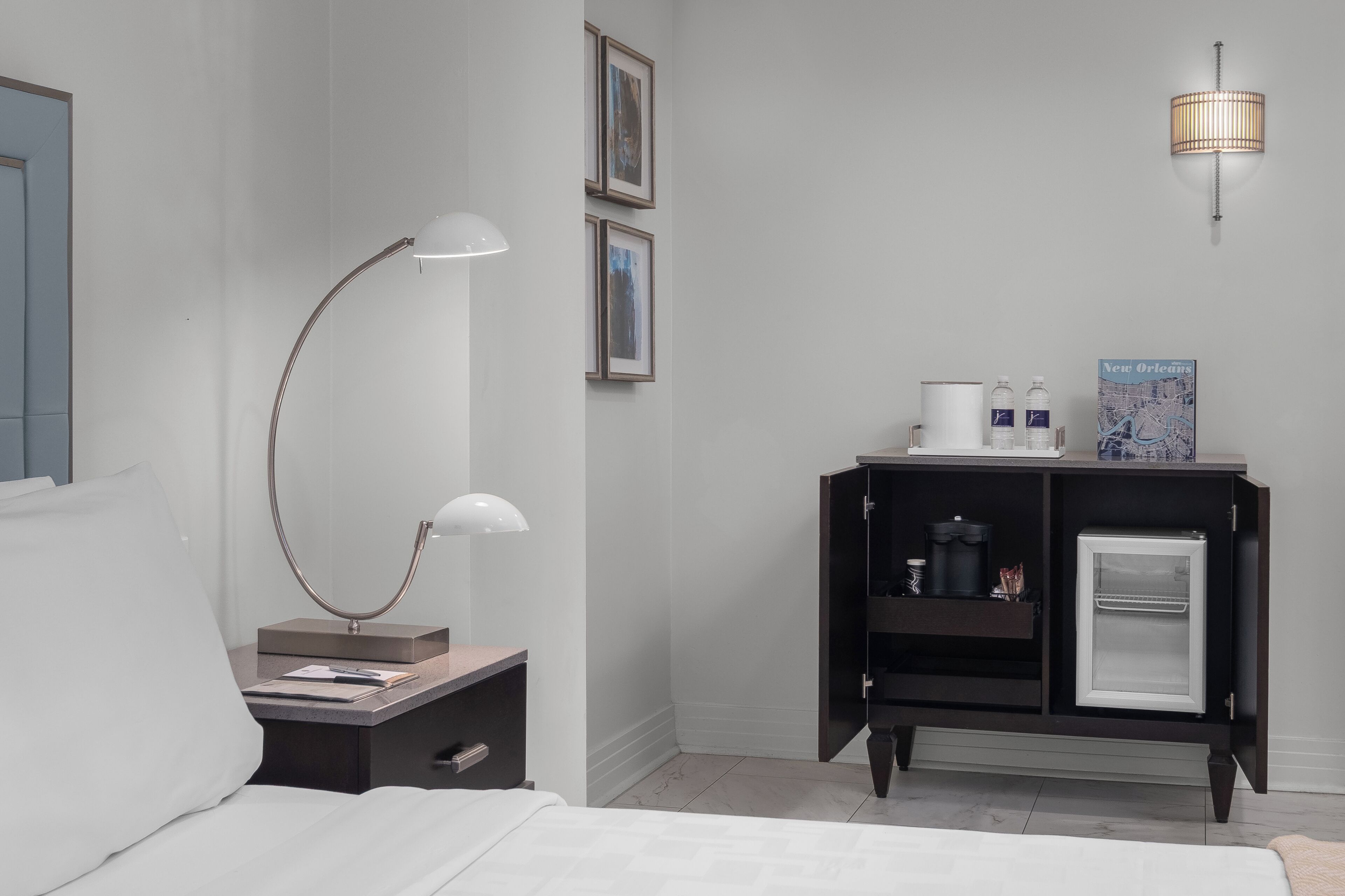 A hotel room with a bed, a desk with a lamp, a minibar, and a wall-mounted light fixture.