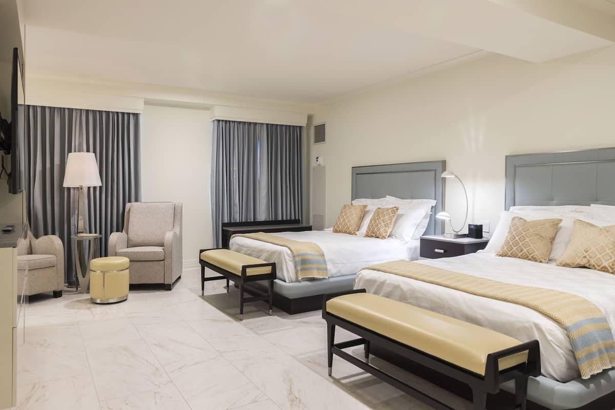 A hotel room with two beds, a sofa, a chair, a bench, a lamp, and a bedside table.