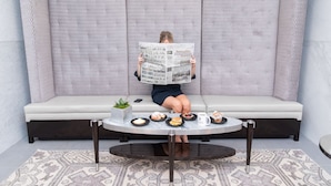 Une personne est assise sur un canapé gris, un journal à la main. Devant elle, il y a une petite table sur laquelle sont disposées des pâtisseries et une tasse de café.