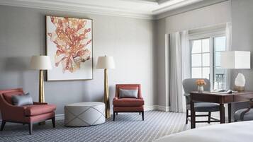 A hotel room with a large bed, two armchairs, a round ottoman, a desk with a chair, and a large abstract painting on the wall.