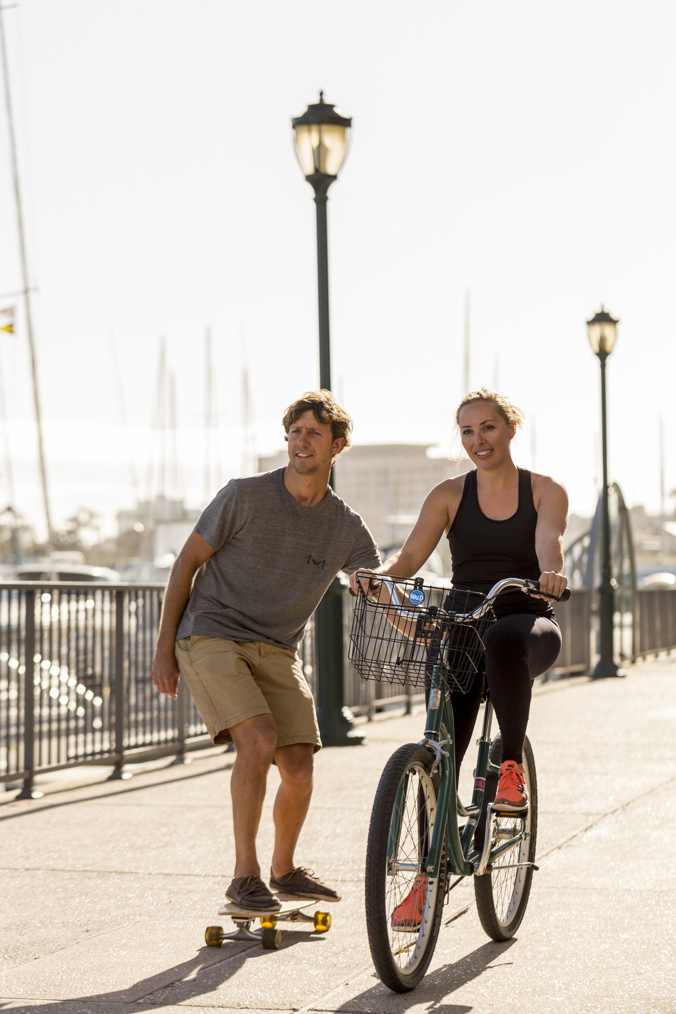 Deux personnes sur un vélo et une personne sur une planche à roulettes, sur un pont.