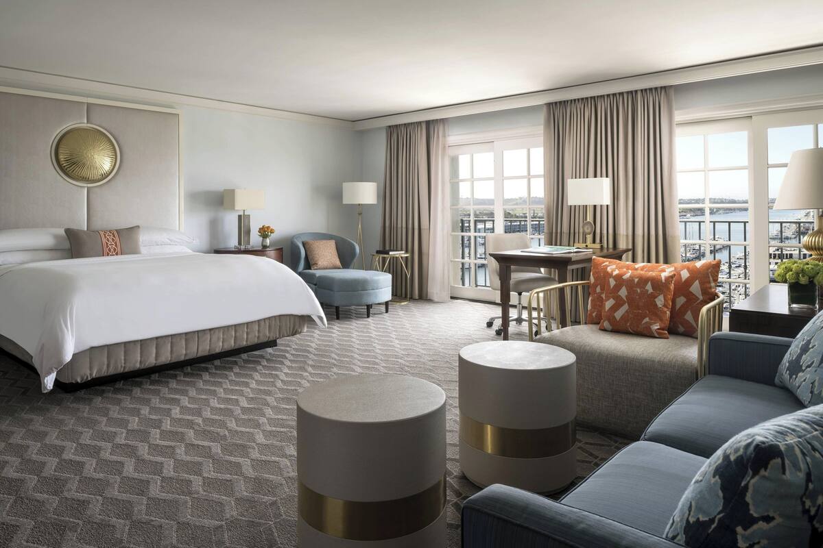 A hotel room with a large bed, a sofa, two round side tables, a chair, a small table, and a view of the city through large windows.