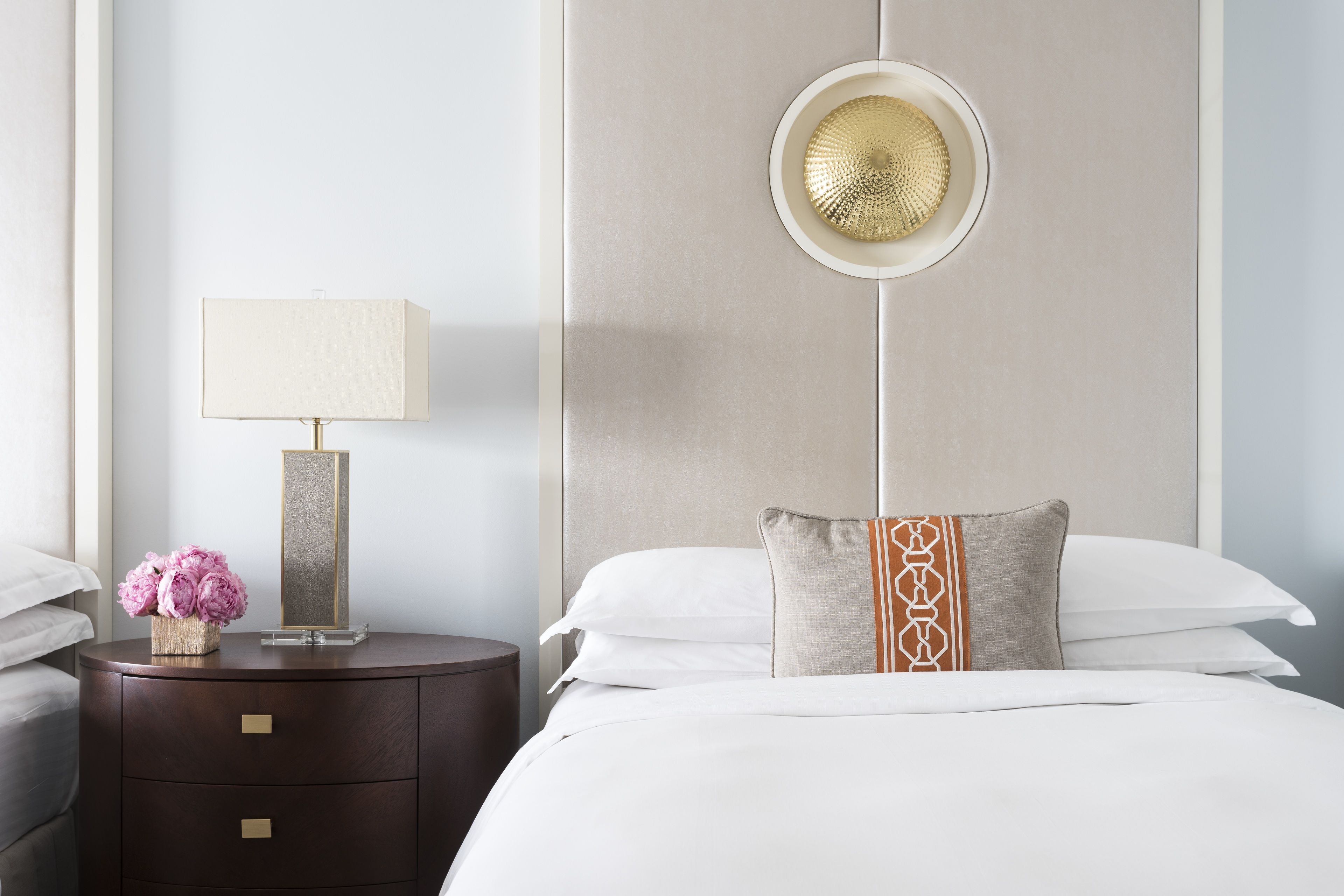 A hotel room with a bed, a bedside table with a lamp, a decorative vase with flowers, and a wall-mounted light fixture.