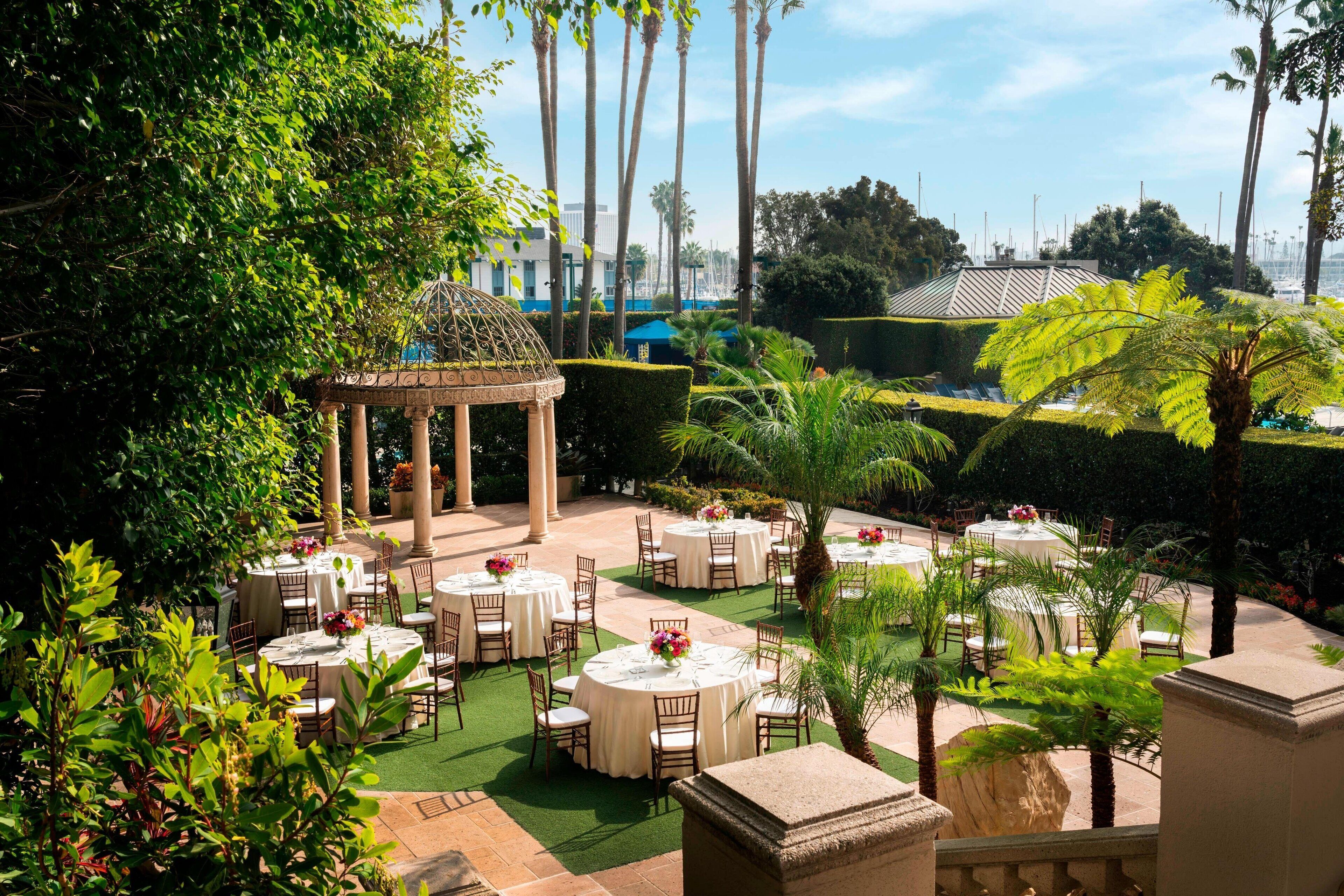 An outdoor dining area with round tables and chairs, surrounded by greenery and palm trees.
