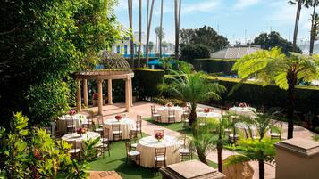 An outdoor dining area with round tables and chairs, surrounded by greenery and palm trees.