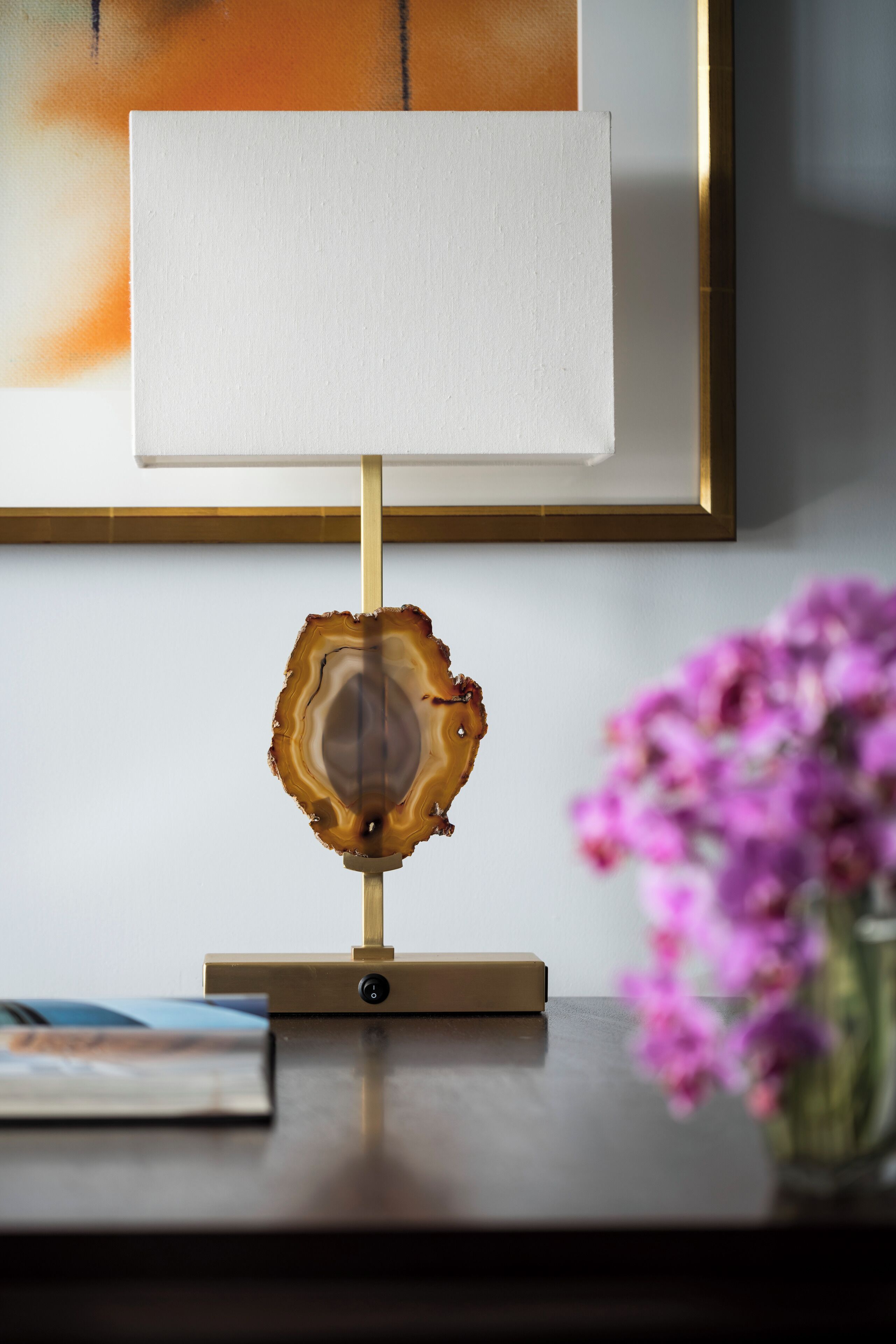 A table with a lamp, a framed artwork, and a vase with flowers.