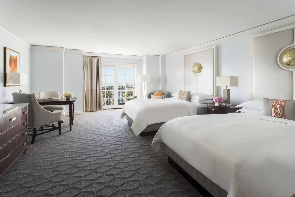 A hotel room with two beds, a desk, a chair, a dresser, and a view of the city through large windows.
