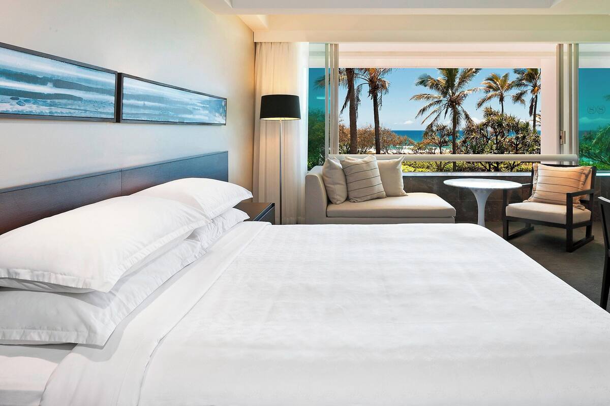 A hotel room with a bed, a sofa, a small table, and a view of palm trees and the ocean.