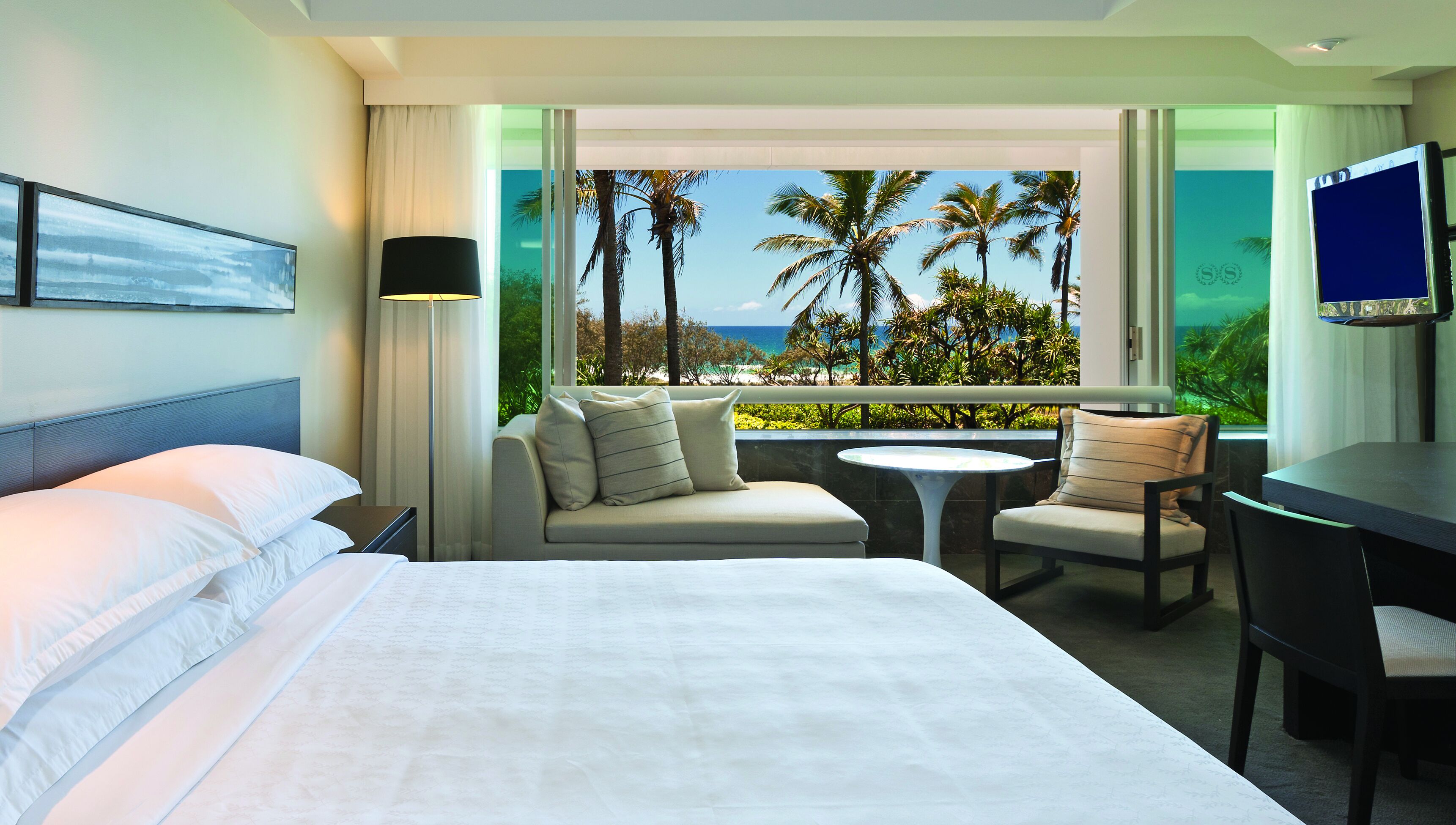 A hotel room with a bed, a sofa, a desk, and a TV. There is a balcony with a view of palm trees and the ocean.