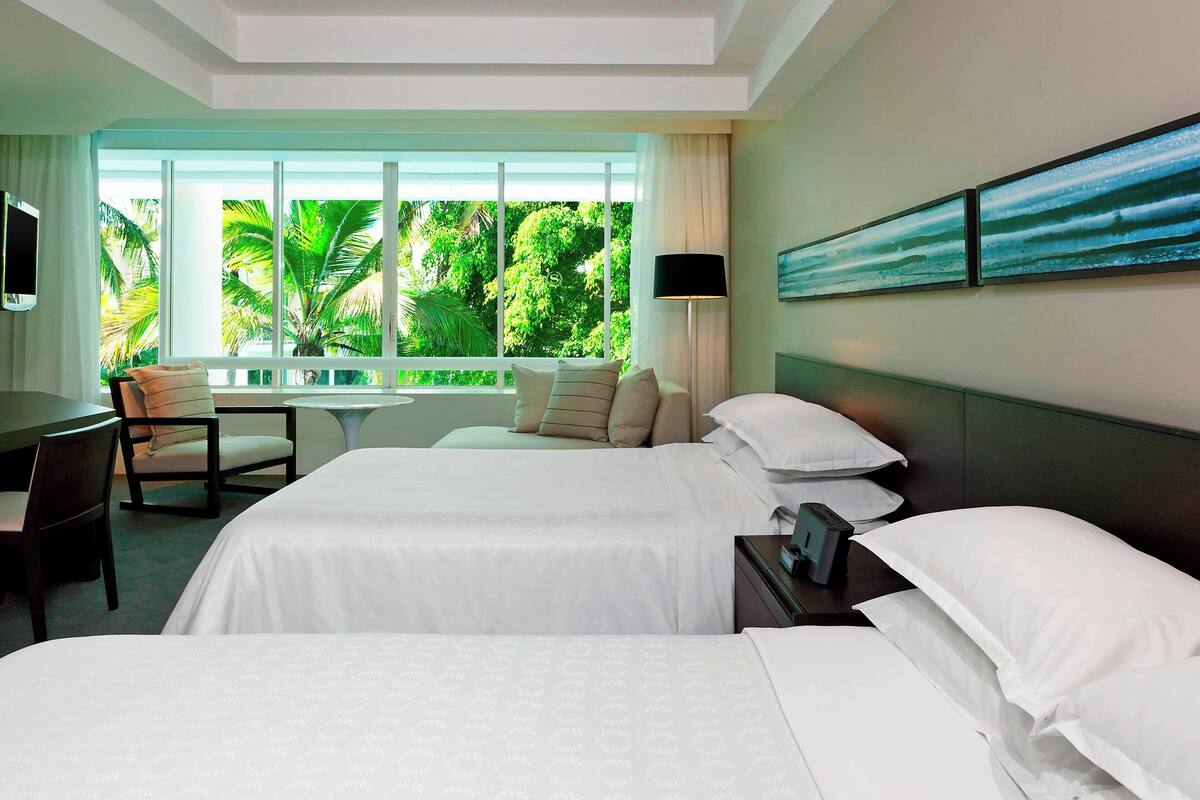 A hotel room with two beds, a desk, a chair, a TV, and a large window with a view of palm trees.