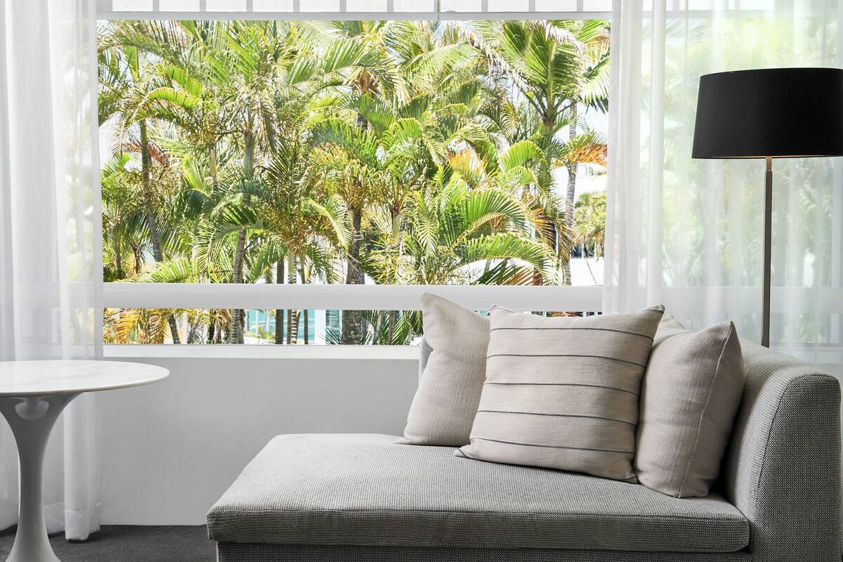 A modern room with a sofa, a small round table, and a view of palm trees through a large window.