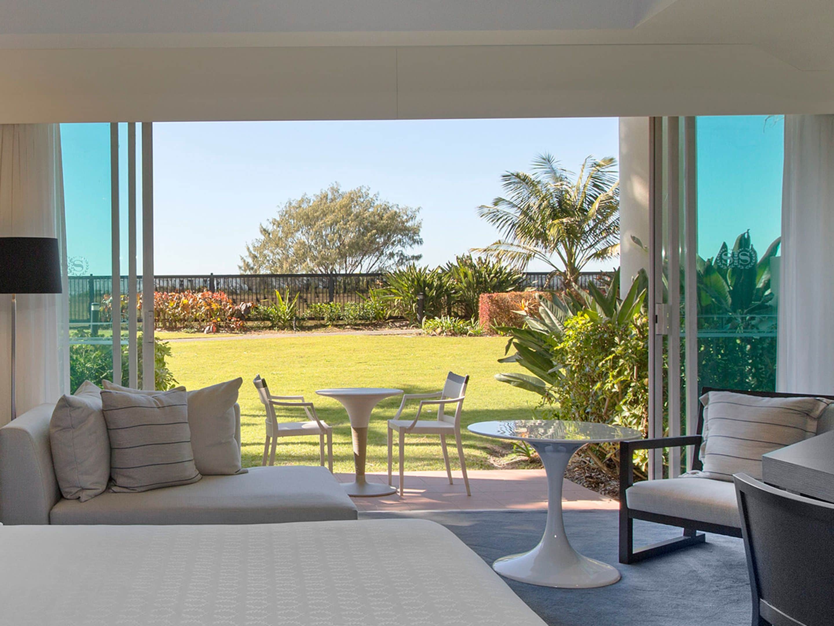 Una habitación de hotel moderna con un balcón grande, una cama, un área de estar con una mesa y sillas, y vistas a un jardín cuidado.
