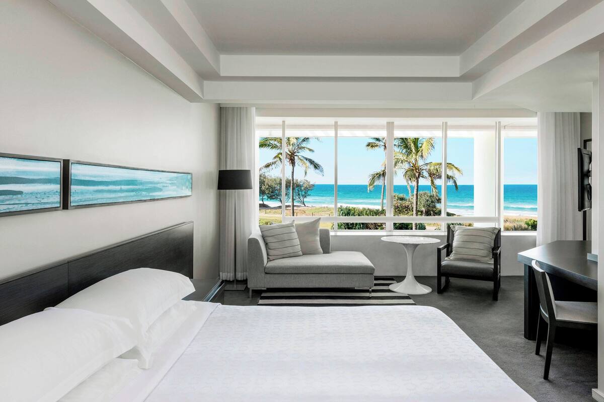 A hotel room with a bed, a sofa, a dining table, and a view of the beach.