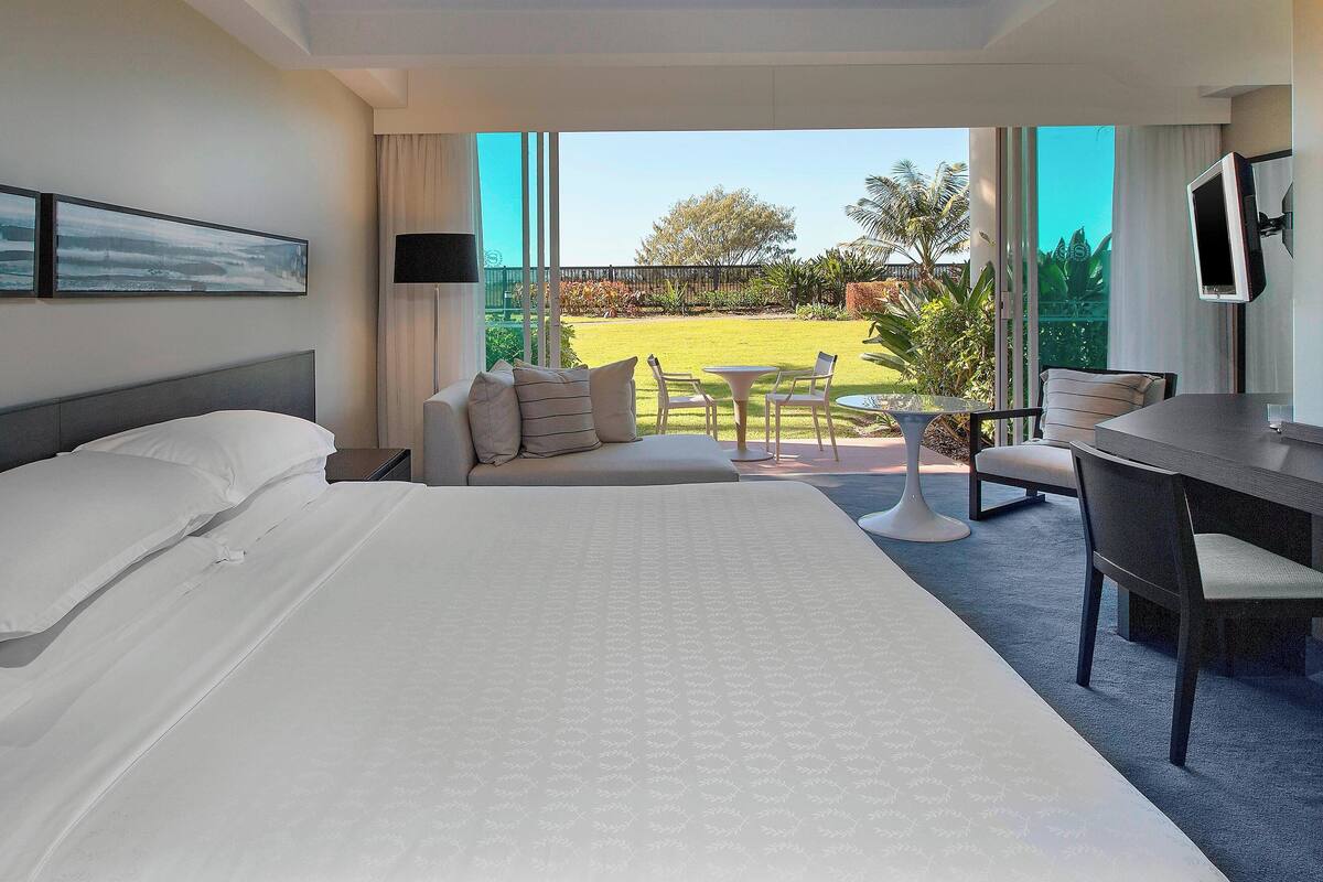 A hotel room with a large bed, a sofa, a small table, and a TV. There is a balcony with a view of the garden and trees.