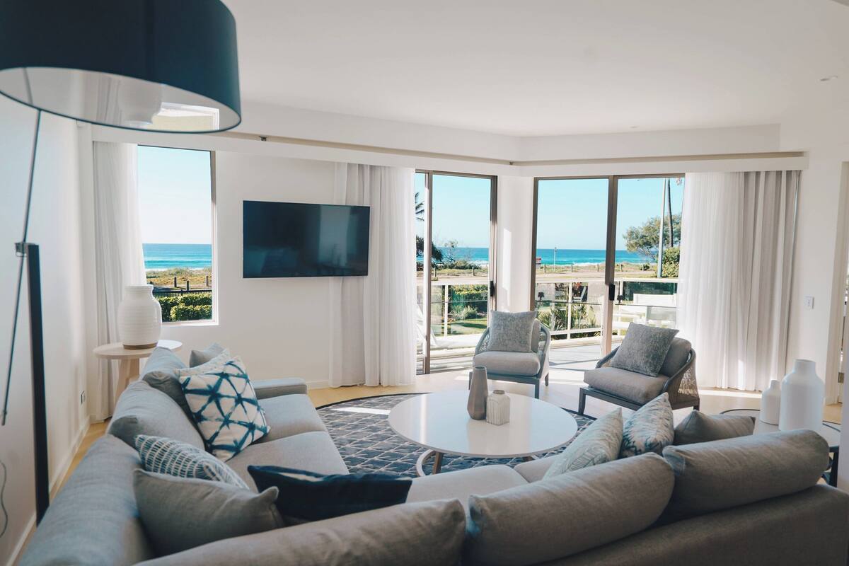 A modern living room with a large sofa, a coffee table, and two armchairs. There is a flat-screen TV mounted on the wall and large windows offering a view of the beach.