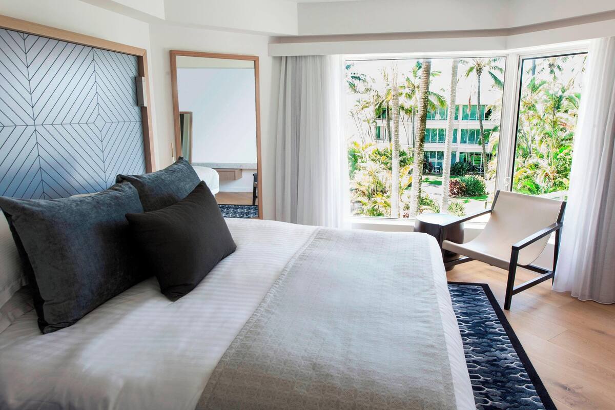 A modern hotel room with a large bed, a view of a tropical garden, and a seating area with a chair and table.