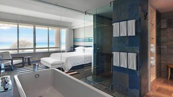 Une chambre d’hôtel moderne dotée d’une grande baignoire, d’une douche en verre, d’un lit avec du linge de maison blanc et offrant une vue sur la mer grâce à de grandes fenêtres.