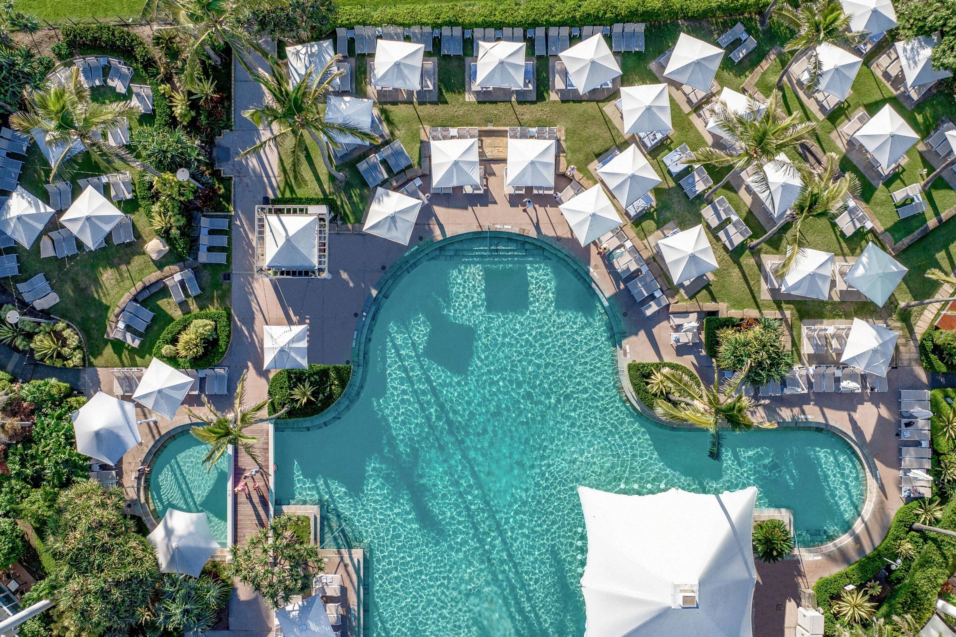 Vista aérea de un resort con piscina central, rodeada de tumbonas y sombrillas, y palmeras.