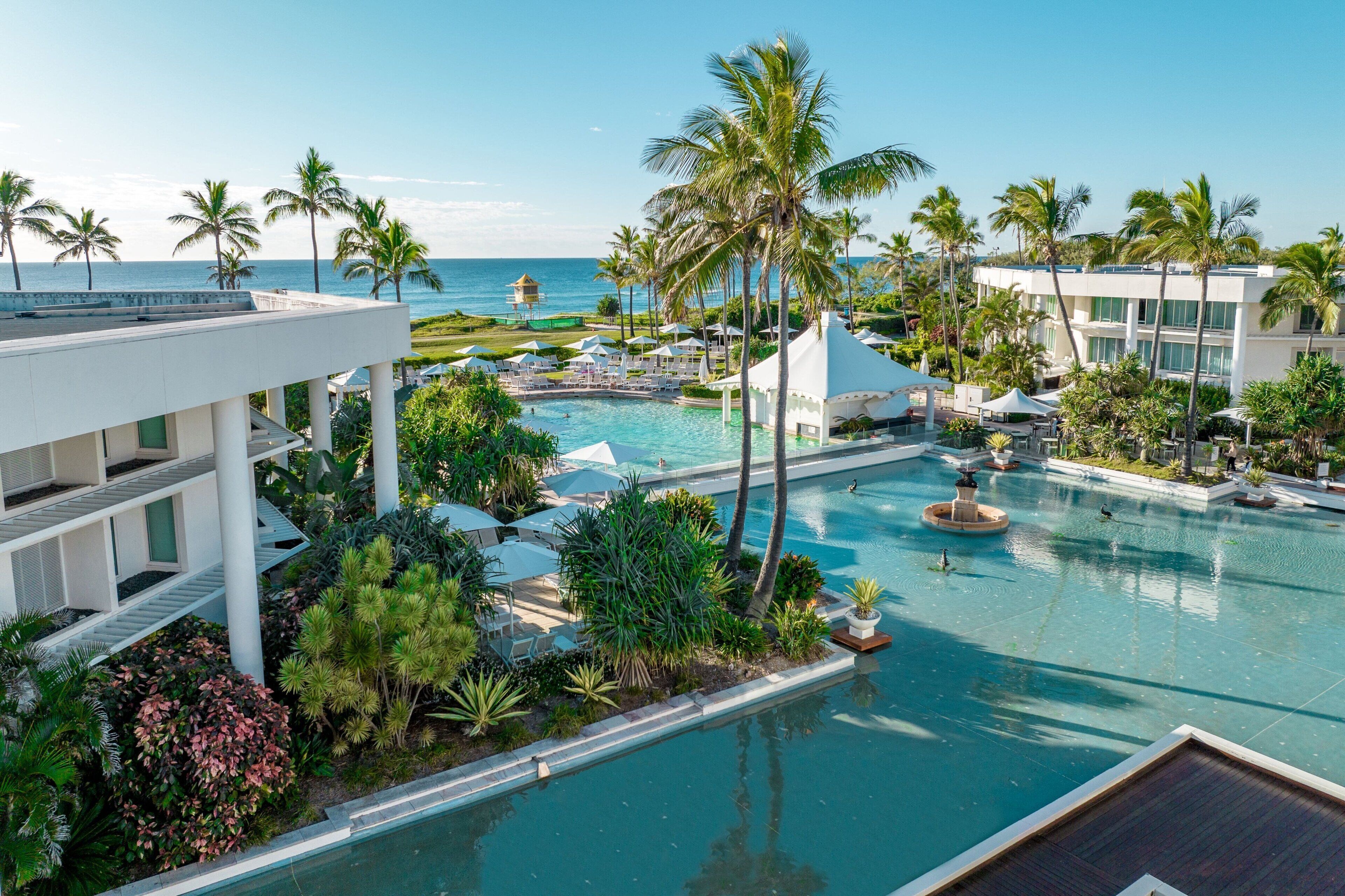 Área de piscina de resort com palmeiras, vista para o oceano e um edifício branco com varandas múltiplas.