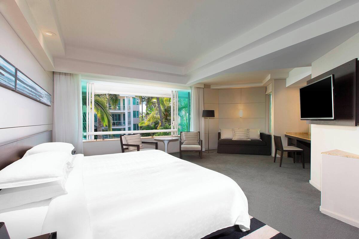 A hotel room with a large bed, a desk, a chair, a TV, and a window with a view of palm trees.