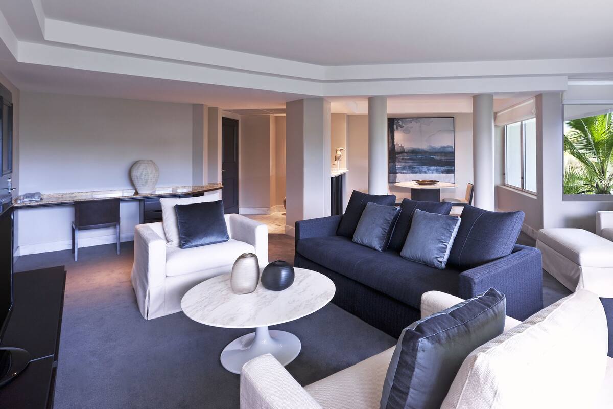 A modern living room with a dark blue sofa, white armchairs, and a round coffee table. A desk and chair are visible in the background, and a painting hangs on the wall.