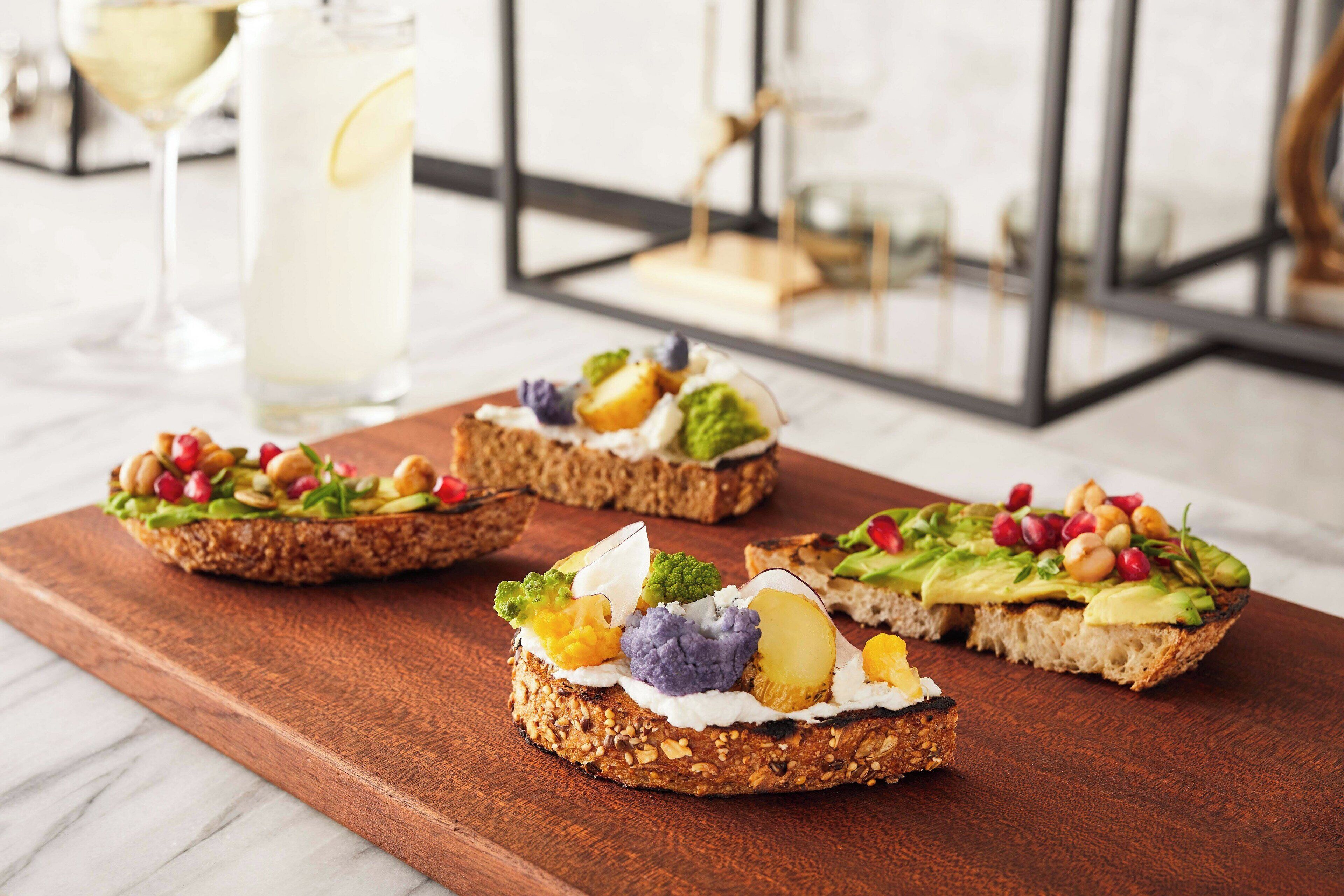Three open-faced sandwiches on a wooden board with various toppings, a glass of a clear beverage, and a glass of a pale beverage in the background.
