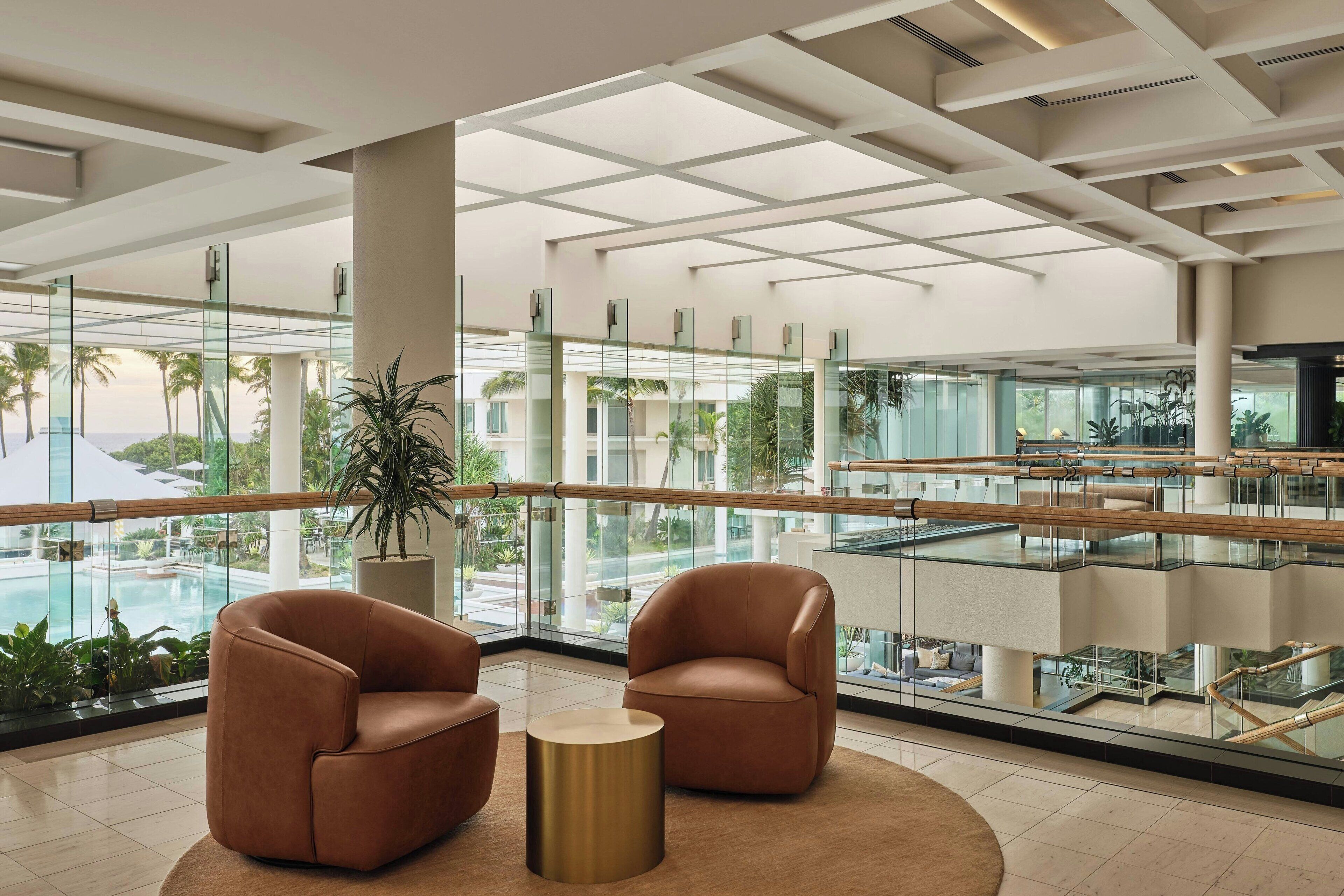 A spacious lobby with a glass railing, two brown armchairs, a round gold table, and a large potted plant.