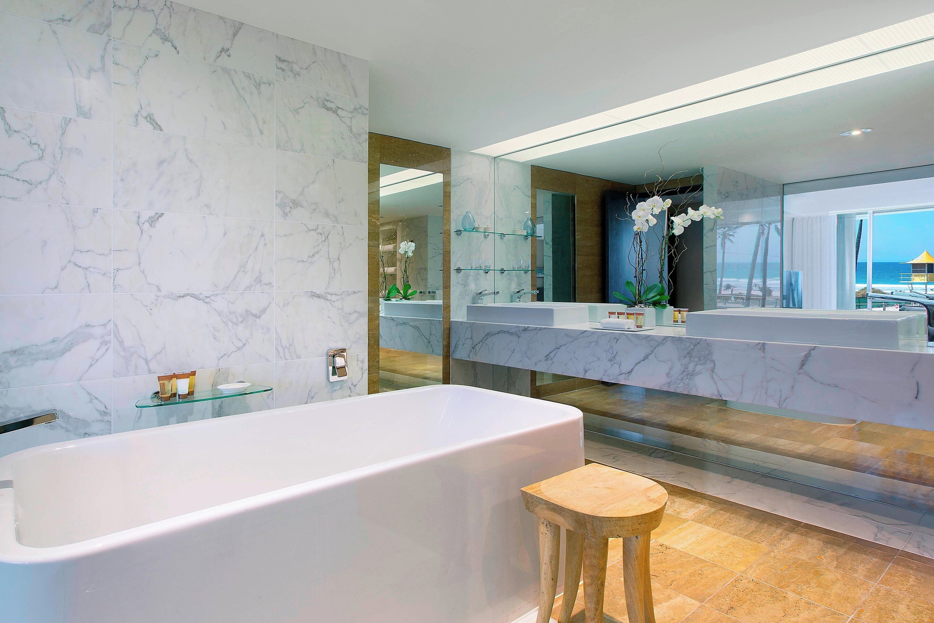 A modern bathroom with a large bathtub, marble walls, a glass shelf, and a wooden stool.