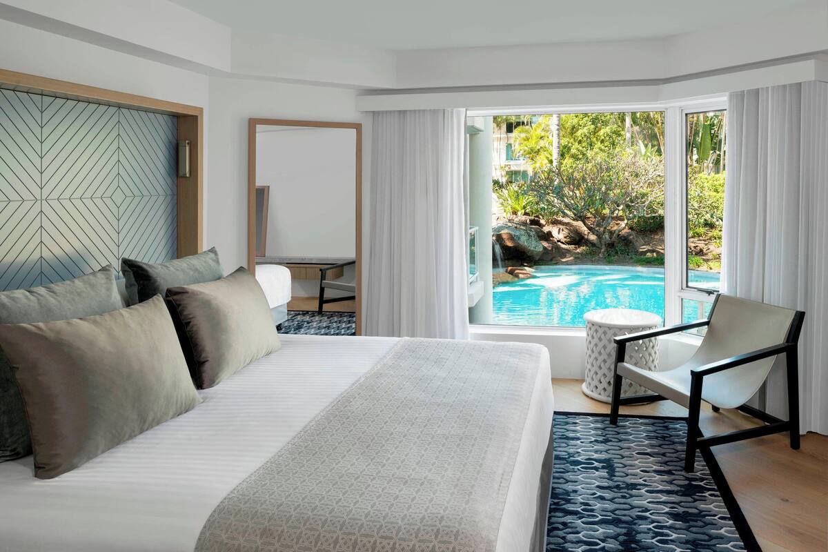 A hotel room with a bed, a chair, a small table, and a view of an outdoor swimming pool.