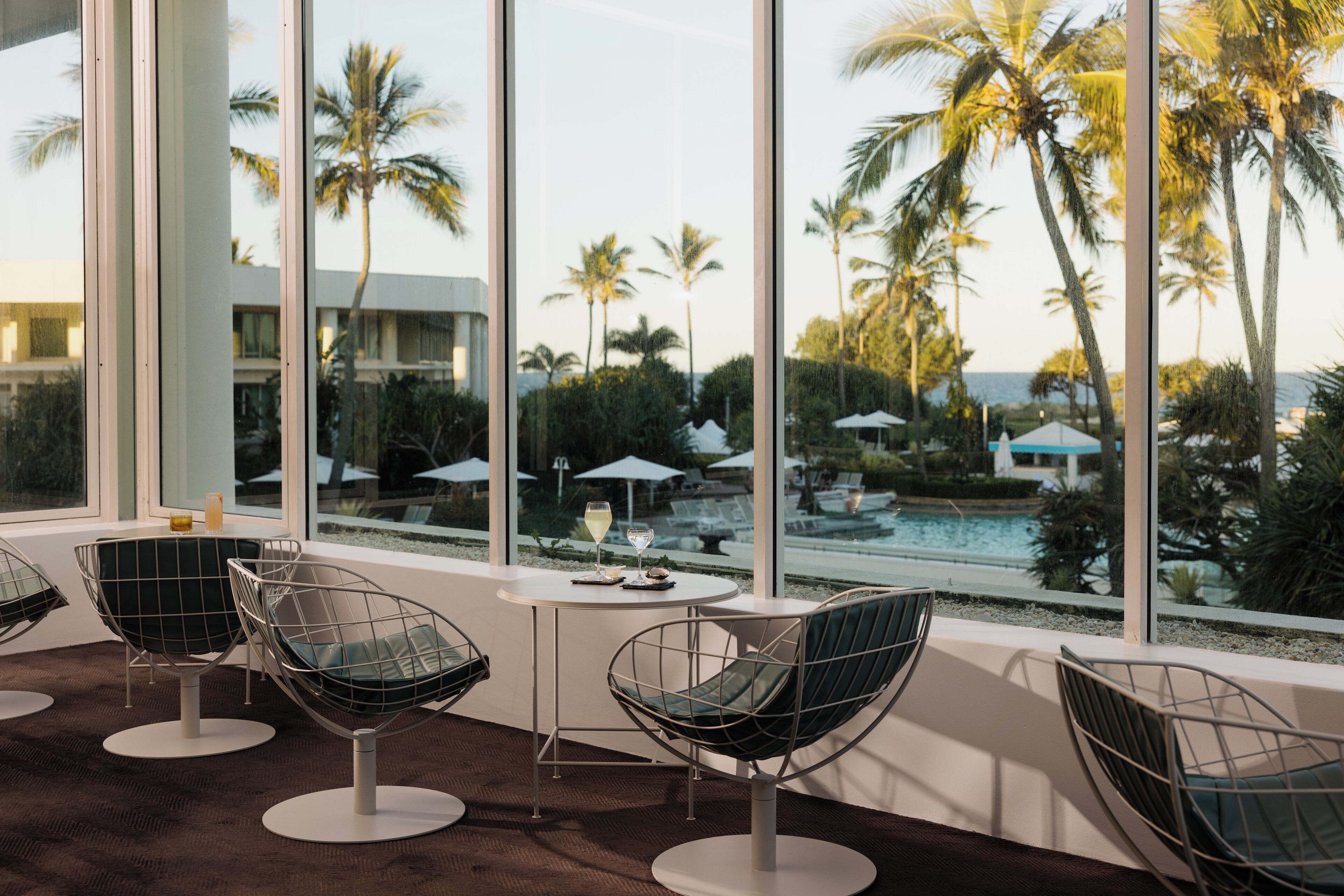 A modern room with large windows overlooking a pool and palm trees. There are three wireframe chairs and a small table with a glass of wine.