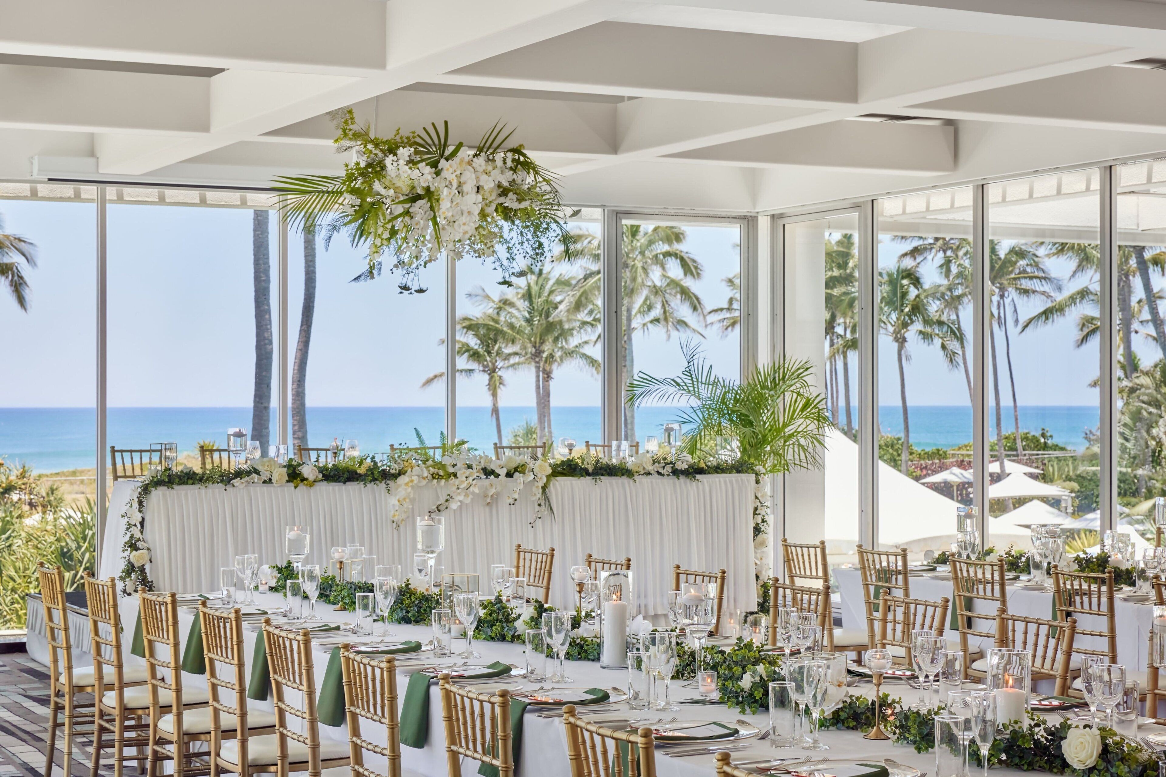 An outdoor dining area with a view of the ocean, featuring a white tablecloth, green napkins, and wooden chairs.