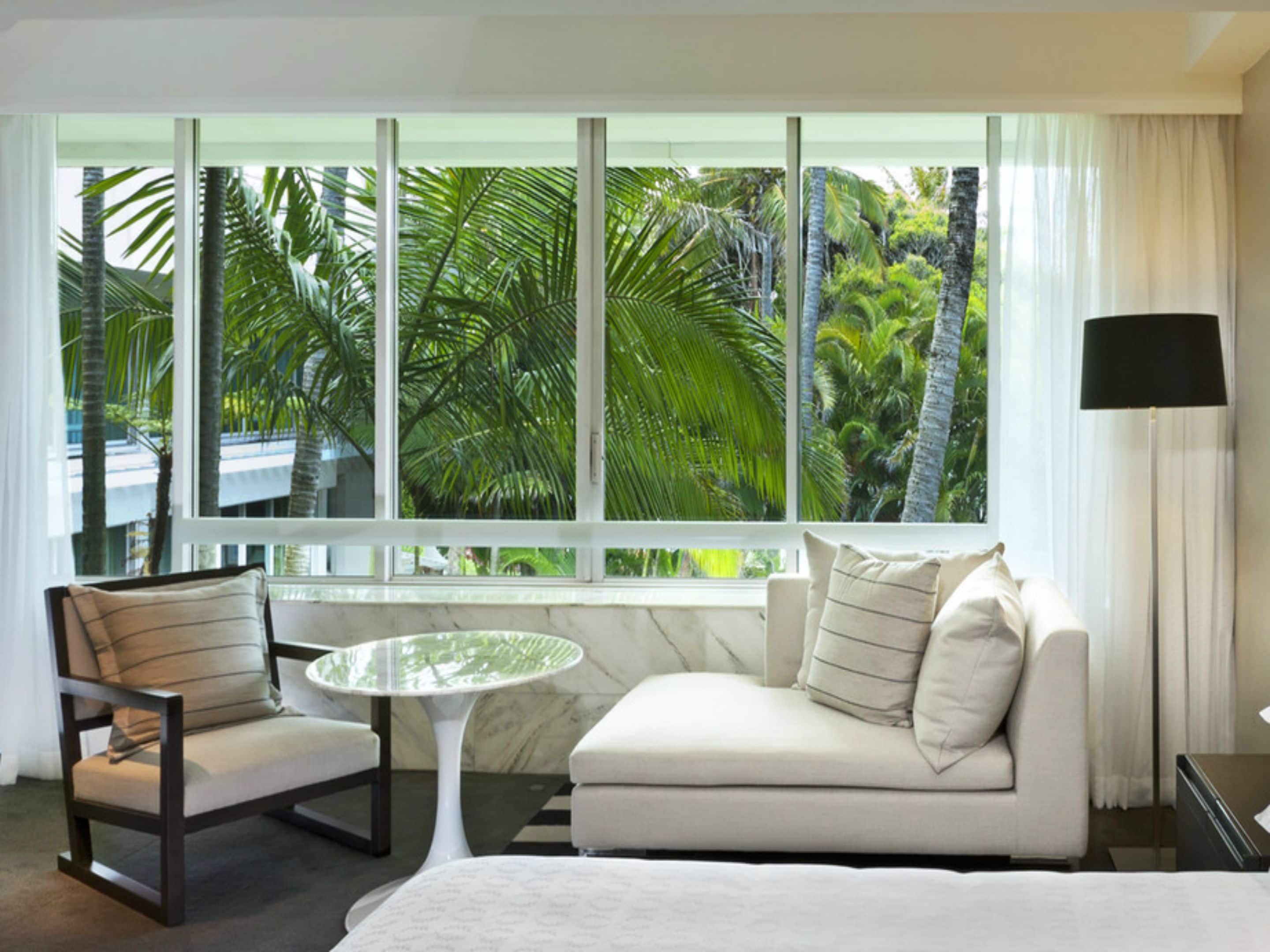 A modern hotel room with a large window, a white sofa, a small round table, and a chair.