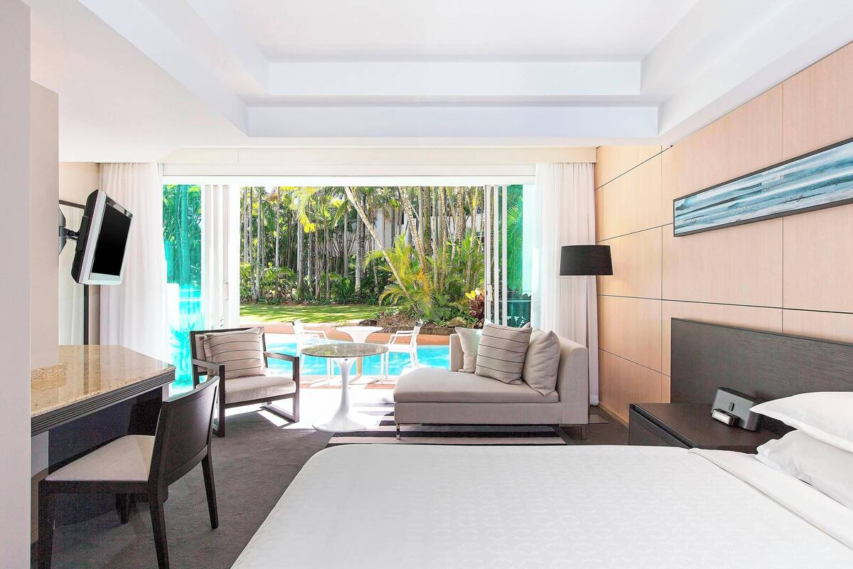 A hotel room with a bed, a desk, a chair, a small table, and a view of a swimming pool and greenery.