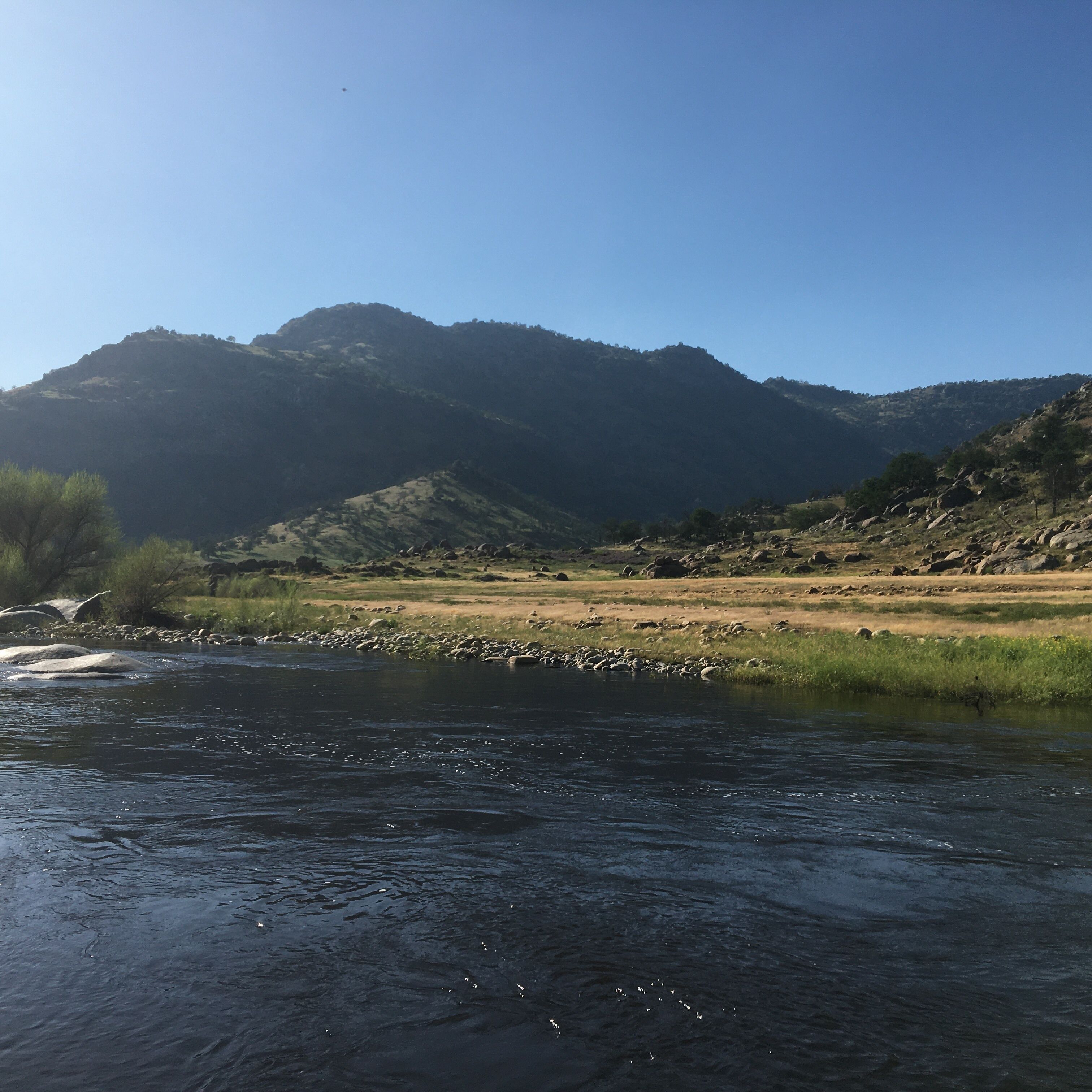 A river with a rocky bed, surrounded by grassy areas and hills.