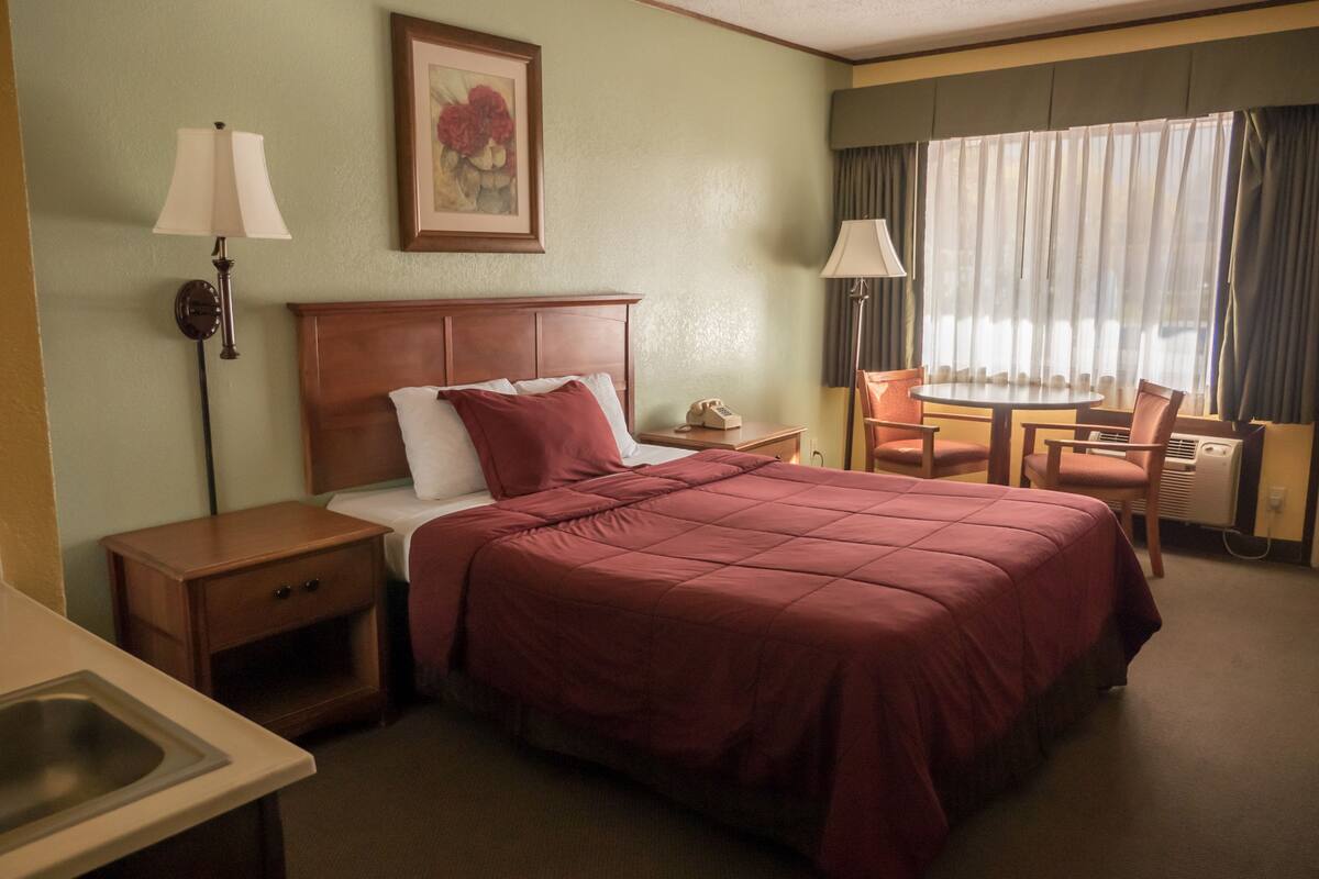 A hotel room with a large bed, a bedside table, a chair, a desk, and a window with curtains.