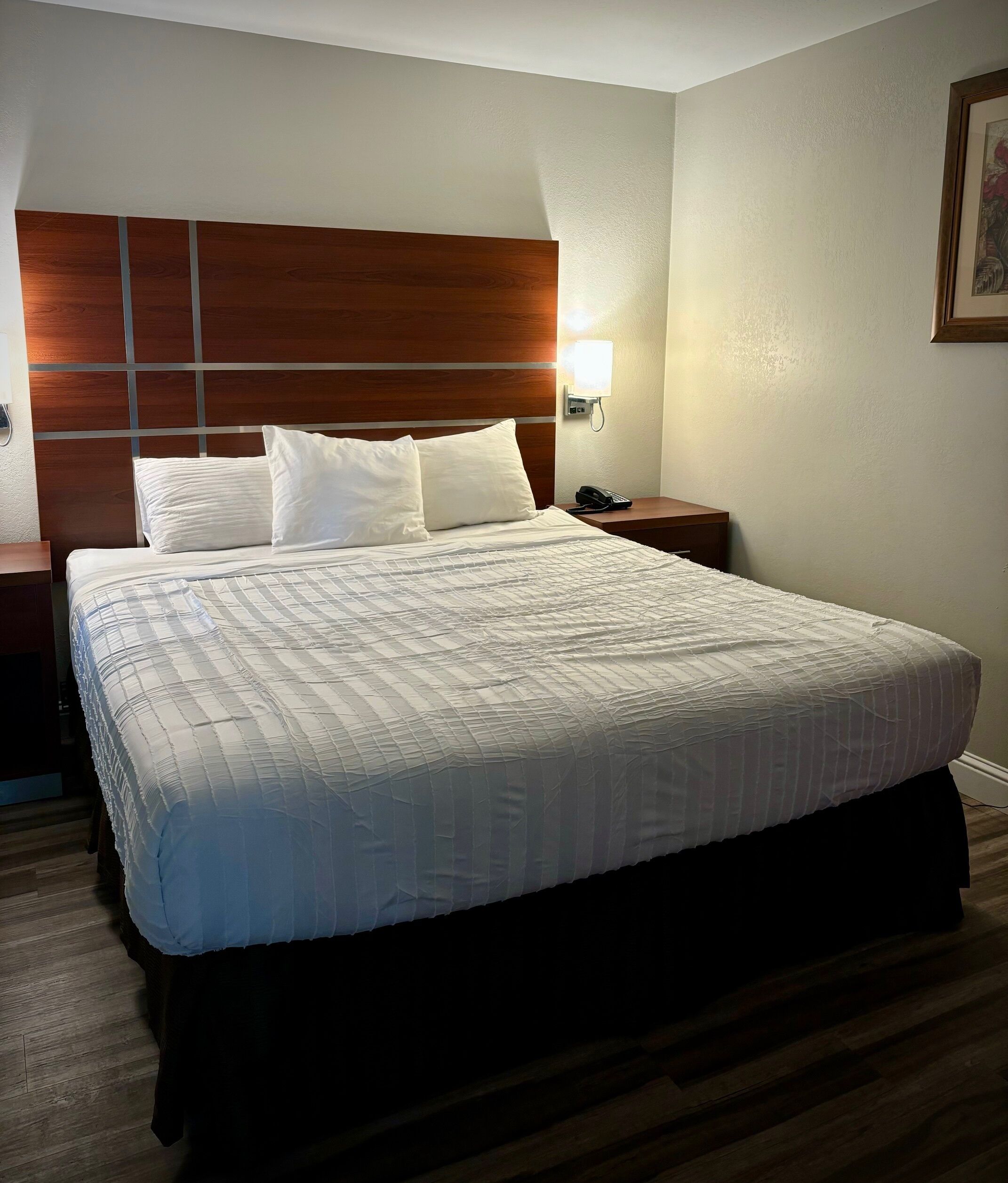 Hotellihuone, jossa on suuri sänky, puinen pääty, yöpöytä lampun kanssa ja seinällä oleva kehystetty kuva.