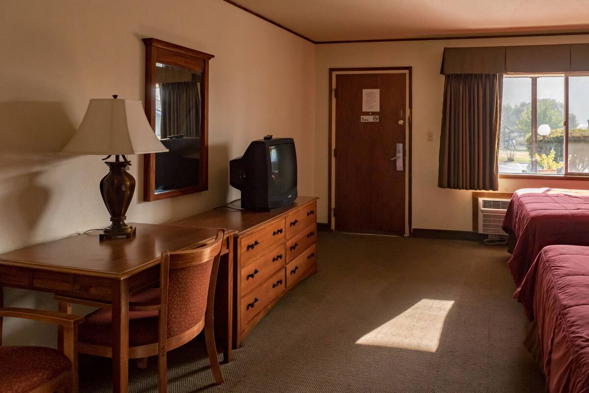 A hotel room with a wooden desk, a chair, a dresser, a television, a mirror, a door, and two beds with red bedding.