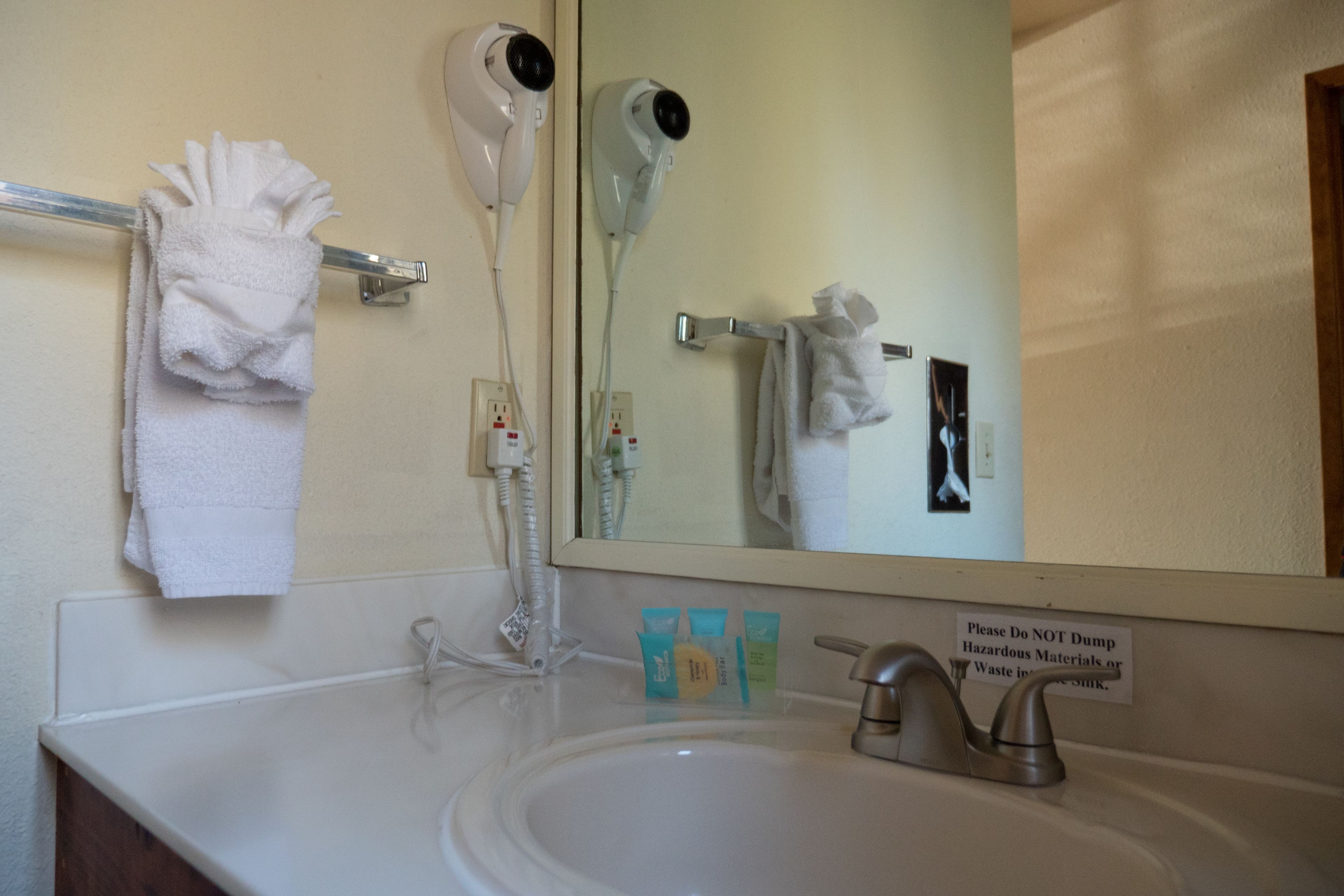 A bathroom with a sink, a mirror, a towel rack, and a hairdryer. There is a sign on the mirror advising not to dump hazardous materials.