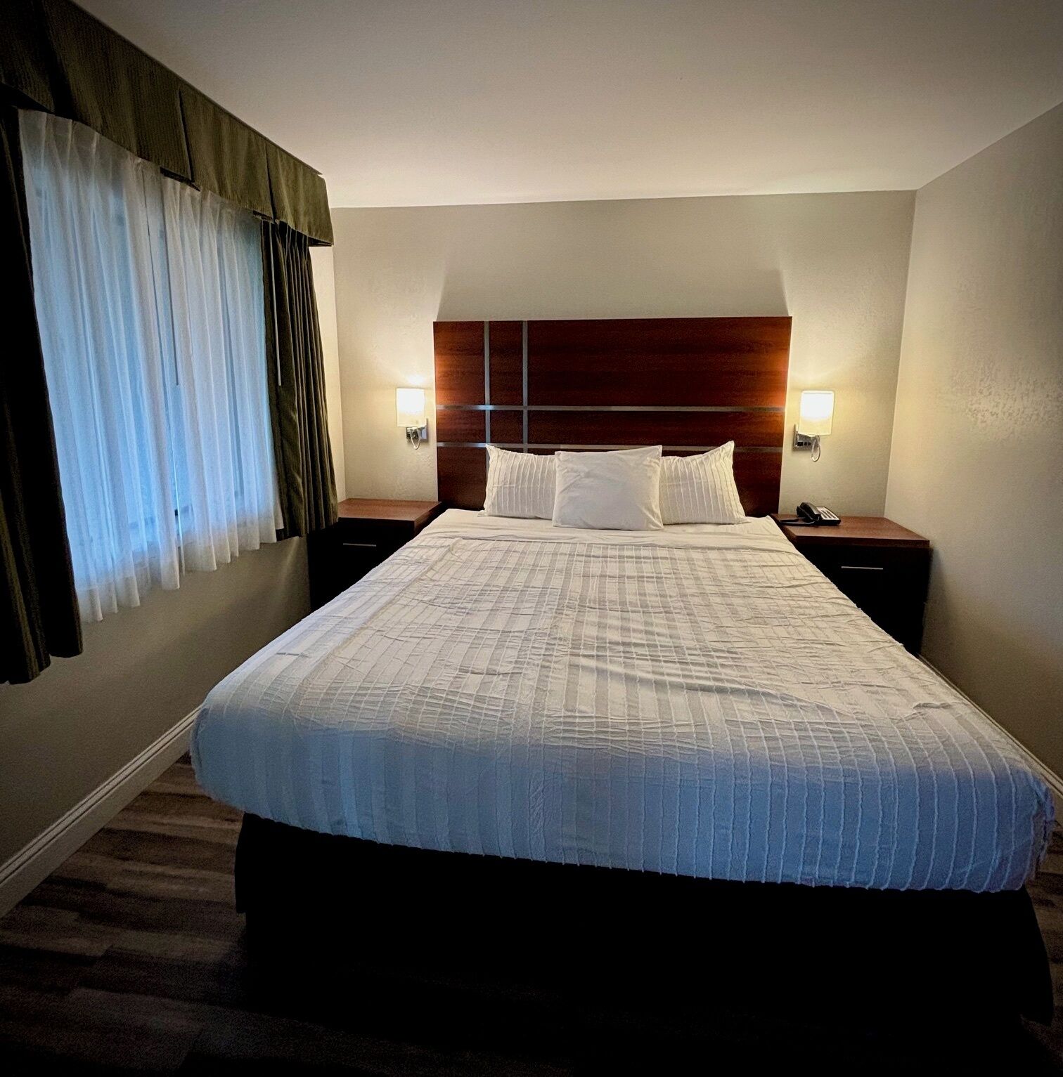 Une chambre d’hôtel avec un grand lit, deux tables de chevet, une tête de lit et une fenêtre avec des rideaux.