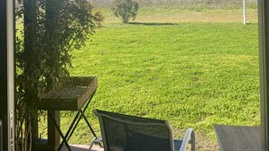 Vista desde una habitación con puerta de vidrio a un área exterior con césped, una pequeña mesa y una silla.