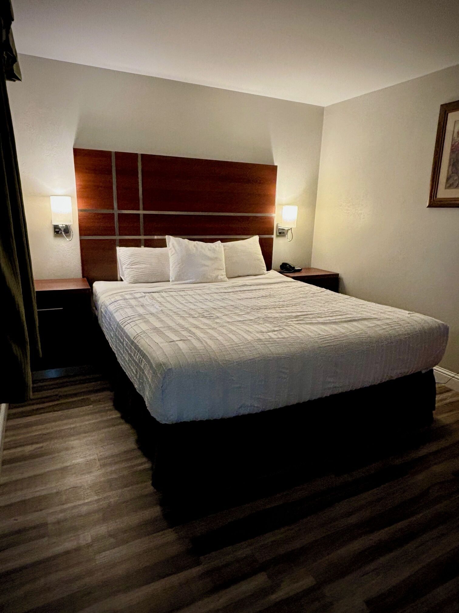 A hotel room with a large bed, wooden headboard, bedside lamps, and a framed picture on the wall.