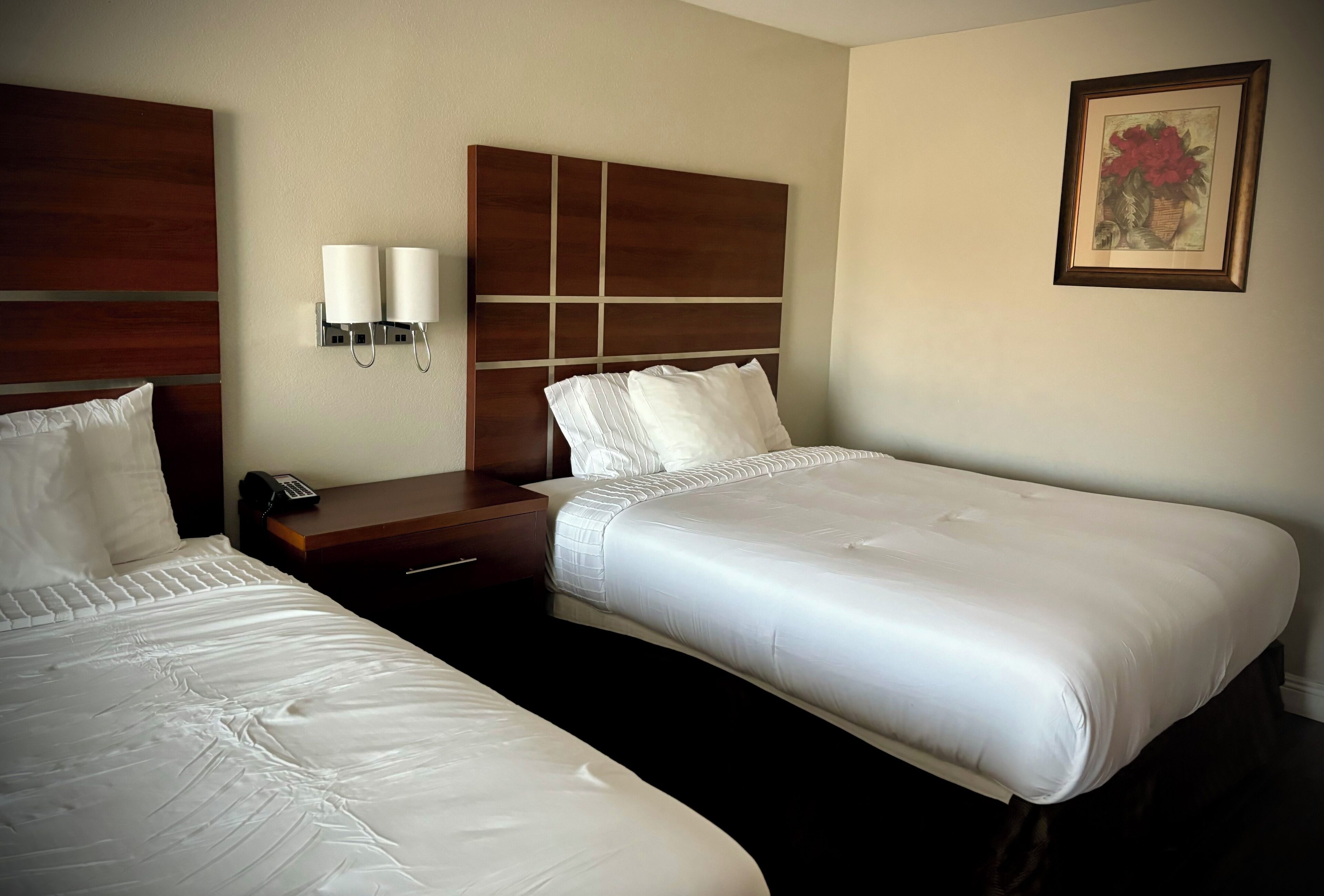 A hotel room with two beds, a wooden headboard, bedside lamps, a nightstand, and a framed picture on the wall.