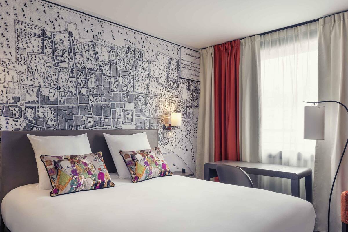 Ein Hotelzimmer mit einem großen Bett, zwei Zierkissen, einem Schreibtisch, einem Sessel, einer Lampe und einem Stadtbild als Wandbild.