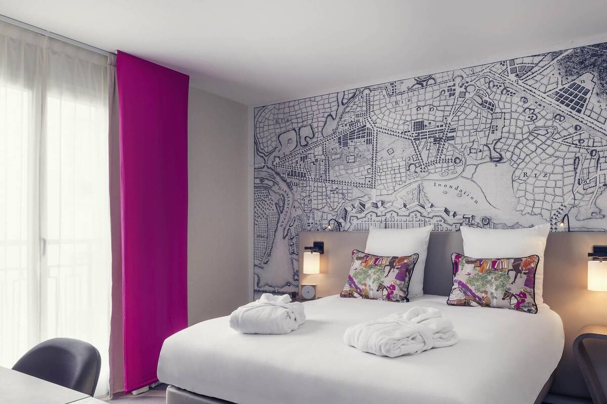 Ein Hotelzimmer mit einem großen Bett, zwei Kissen mit bunten Mustern, einer Karte an der Wand, einem Schreibtisch und einem Stuhl.